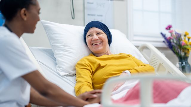 A female nurse sits at the bedside of a mature female oncology patient as she checks in on her during rounds. The patient is dressed comfortably and has a head scarf on to keep warm.
