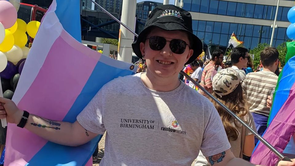 Avery Cunningham stands wearing a bucket hat and sunglasses and holding a transgender pride flag.