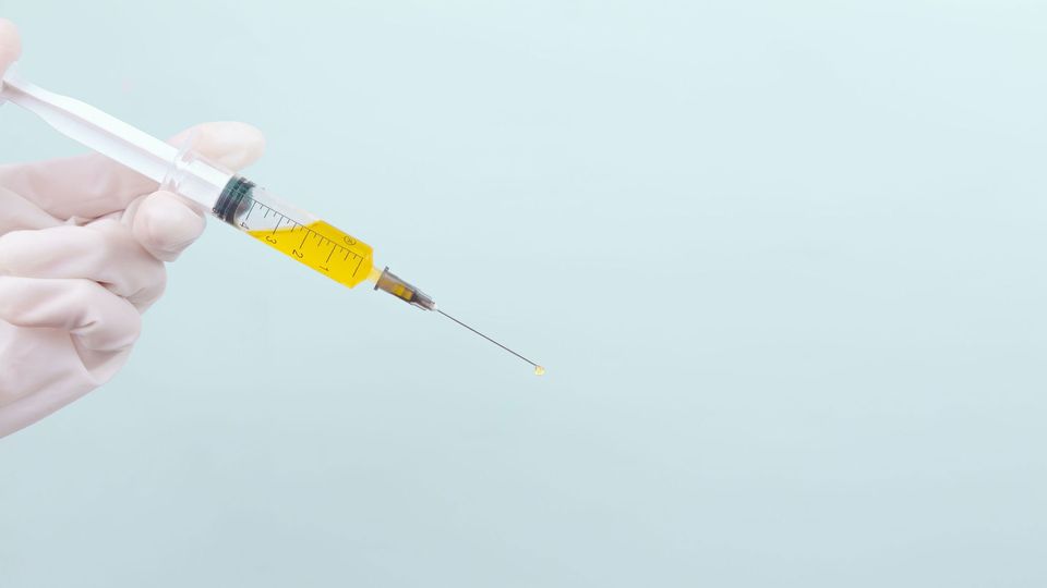 A hand wearing a white glove holds a syringe filled with yellow liquid.