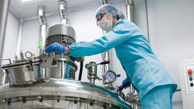 A lab technician in protective gear operates a large stainless steel bioreactor in a cleanroom, adjusting its components