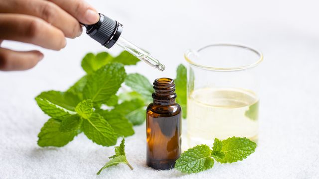 Glass pipette removing peppermint oil from a brown glass bottle next to sprigs of mint leaves and a beaker containing peppermint oil.