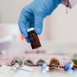 A close-up of a gloved hand holding a small glass vial, with several other vials scattered in the background. The vials are stored in a drawer or tray, suggesting a laboratory or medical setting. 