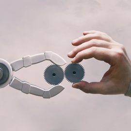 A robot arm holding a gear touching a human hand also holding a gear 